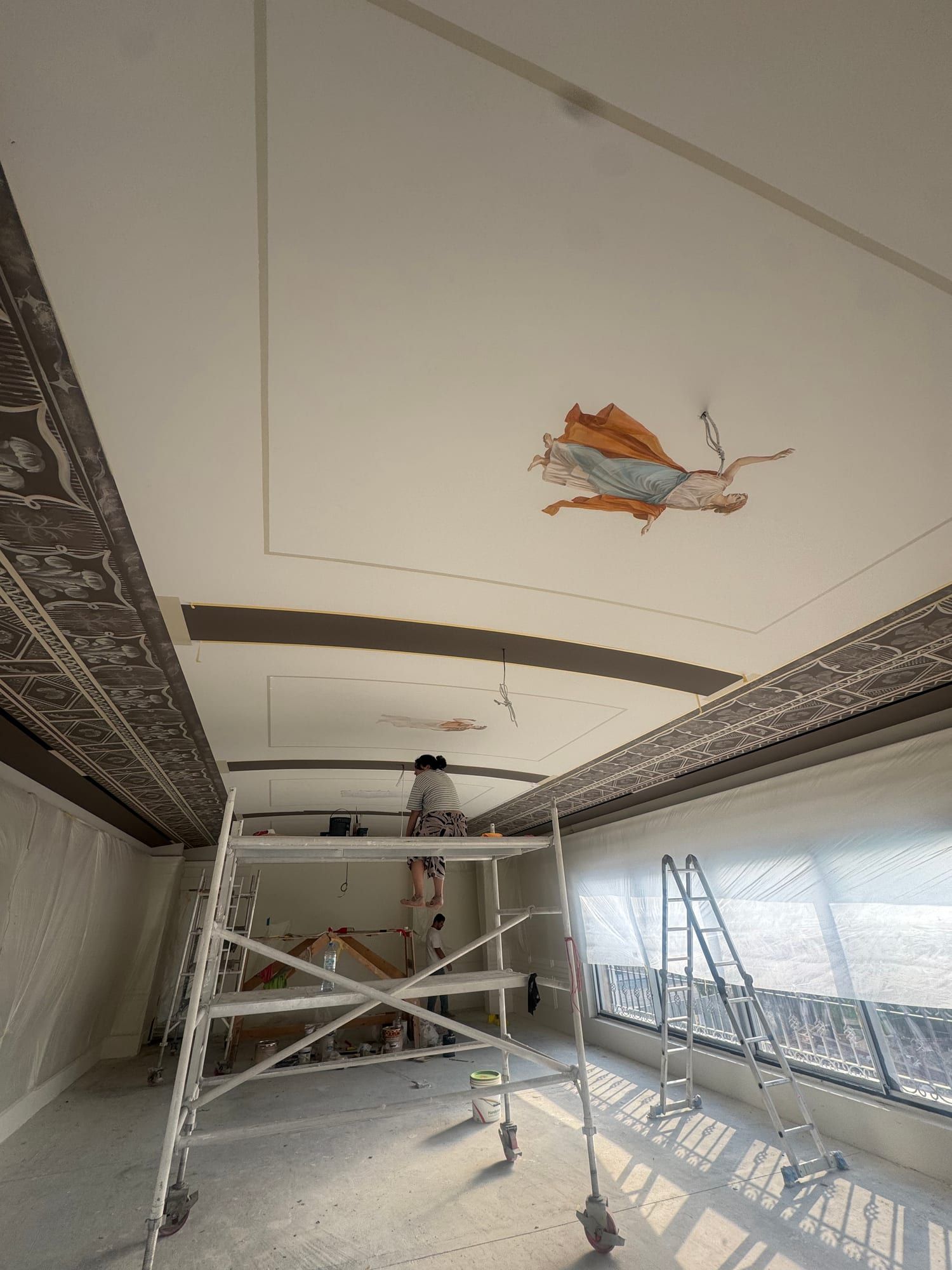 Artist on a mobile scaffold painting a figure onto the ceiling of a long room
