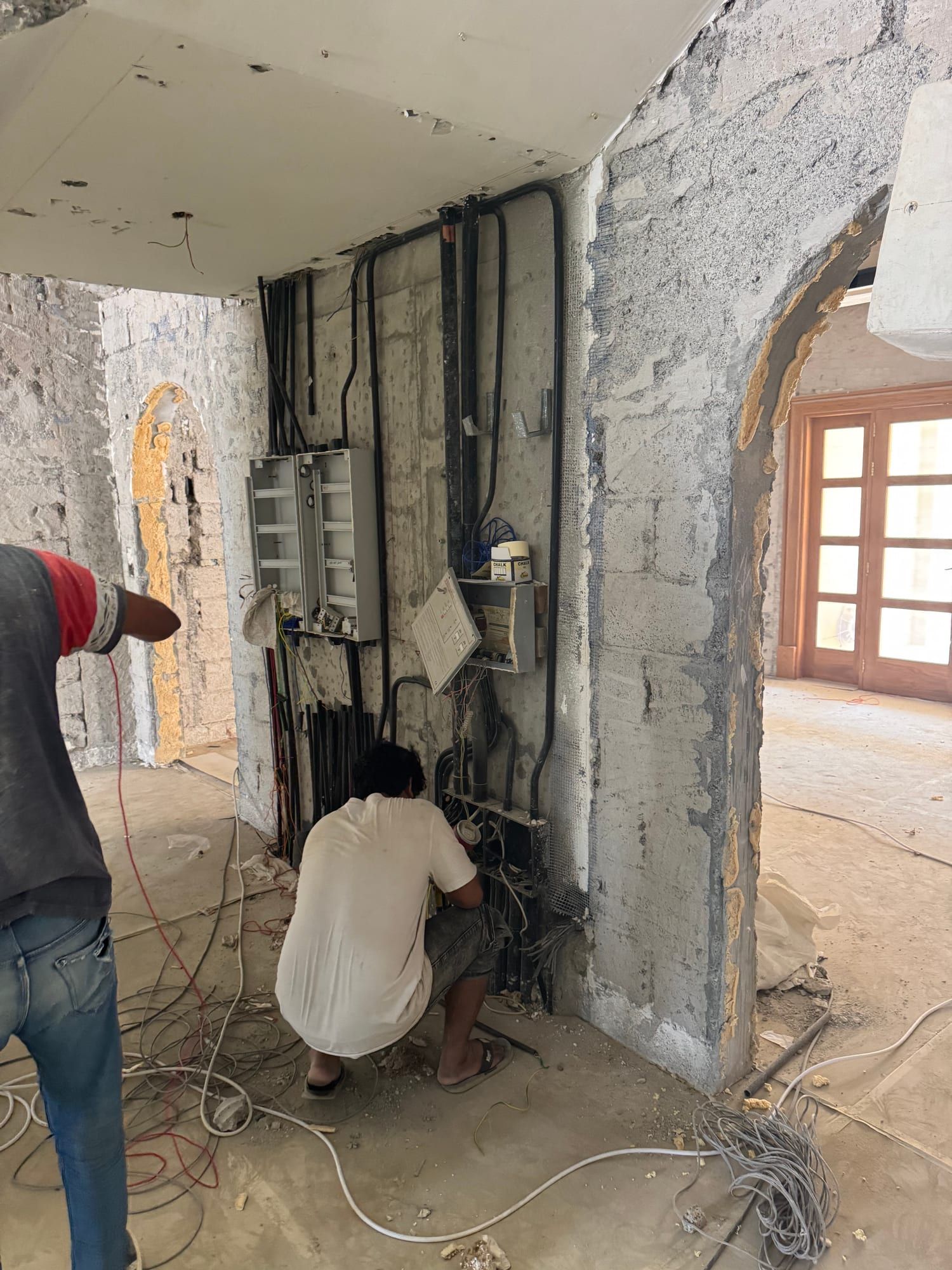 Two electricians working at a vertical run of new conduits and distribution boxes on a stripped wall
